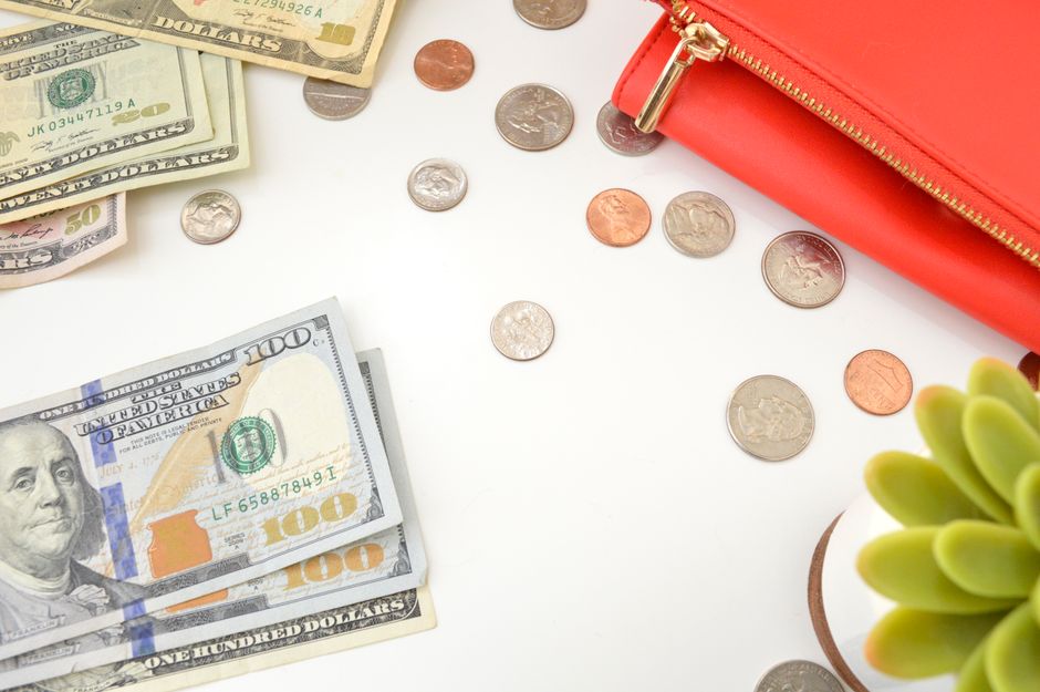 A top view of US dollar bills and coins scattered with a red wallet and succulent plant.