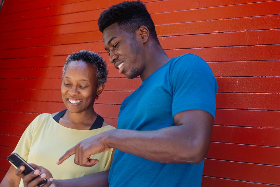 A cheerful mother and son are smiling while looking at a smartphone against a red wall outdoors in daylight.
