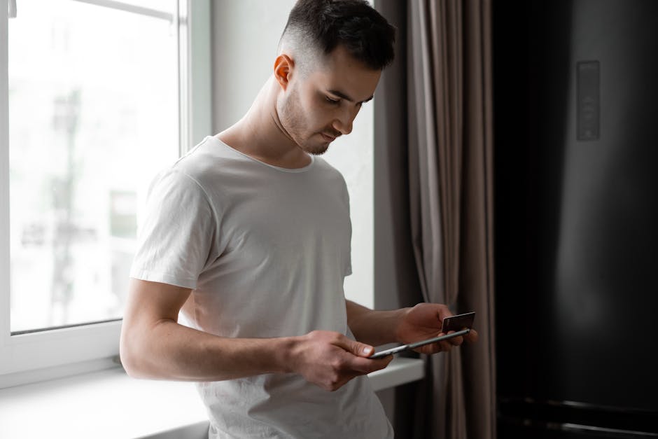 Young man standing by a window, using tablet and credit card for online shopping indoors.