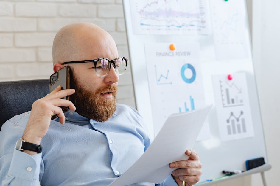 Bald man with beard holding smartphone and papers, reviewing financial charts in office.