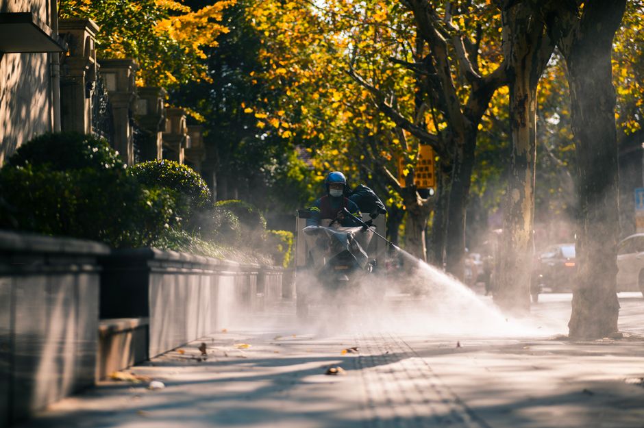 A city worker sprays a sidewalk with water on a sunny day, surrounded by lush trees and buildings.